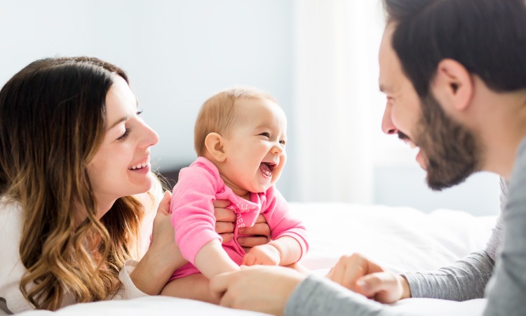 A mom and dad smile at their young baby girl wearing a pink onesie while on a bed